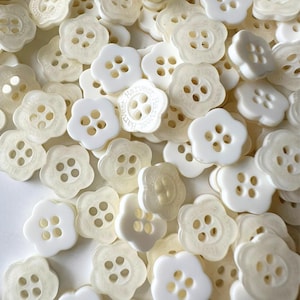 May include: A pile of white plastic flower-shaped buttons with four holes each. The buttons have a slightly translucent appearance and some have the text "Harvest  Button" printed on them.