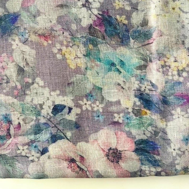 May include: Floral fabric with a watercolor effect. The design features a variety of flowers in shades of pink, white, yellow, and blue, set against a muted purple background. The fabric has a linen-like texture.