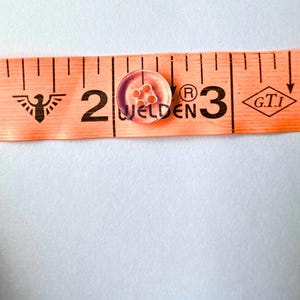 May include: A small, round, clear button with four holes in the centre, positioned on an orange measuring tape that shows the button has a diameter of approximately 1.3 cm. The tape features the text WELDEN, a registered trademark symbol, and G.T.I.