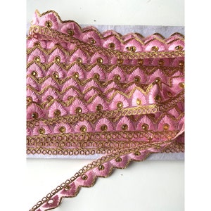 May include: Pink embroidered lace trim with gold accents and small, round, gold-colored embellishments. The trim features a scalloped edge and a repeating geometric pattern. This decorative trim is suitable for various craft projects.