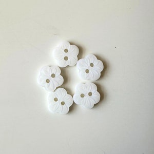 K&ouml;nnte beinhalten: Sechs wei&szlig;e, blumenf&ouml;rmige Kn&ouml;pfe mit jeweils zwei L&ouml;chern. Die Kn&ouml;pfe haben ein erhabenes Design und sind in einem Kreis angeordnet.