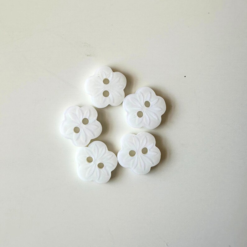K&ouml;nnte beinhalten: Sechs wei&szlig;e, blumenf&ouml;rmige Kn&ouml;pfe mit jeweils zwei L&ouml;chern. Die Kn&ouml;pfe haben ein erhabenes Design und sind in einem Kreis angeordnet.