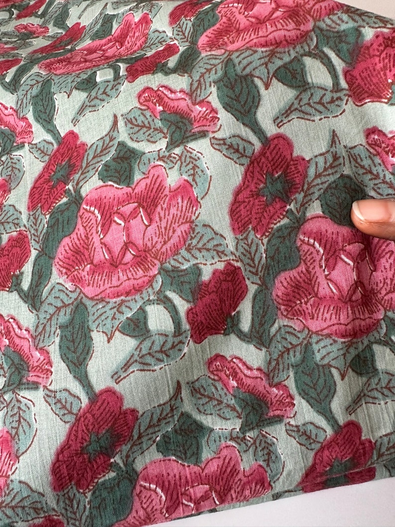 May include: A close-up of a fabric with a floral print. The fabric is a light green color with pink flowers and dark green leaves.