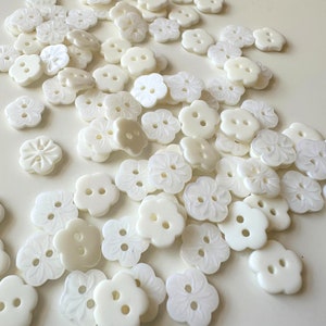 K&ouml;nnte beinhalten: Eine Sammlung wei&szlig;er, perlmuttartiger Kn&ouml;pfe mit Blumenmuster. Die Kn&ouml;pfe haben zwei L&ouml;cher und sind in einem zuf&auml;lligen Muster angeordnet.
