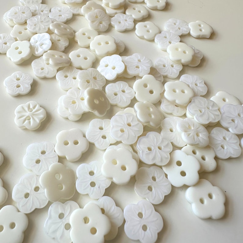 K&ouml;nnte beinhalten: Eine Sammlung wei&szlig;er, perlmuttartiger Kn&ouml;pfe mit Blumenmuster. Die Kn&ouml;pfe haben zwei L&ouml;cher und sind in einem zuf&auml;lligen Muster angeordnet.