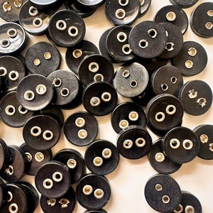 K&ouml;nnte beinhalten: Eine Ansammlung schwarzer, runder Kn&ouml;pfe mit zwei L&ouml;chern und silbernen Metall&ouml;sen. Die Kn&ouml;pfe sind auf einer hellen Oberfl&auml;che verstreut. Geeignet f&uuml;r N&auml;h- oder Bastelprojekte.