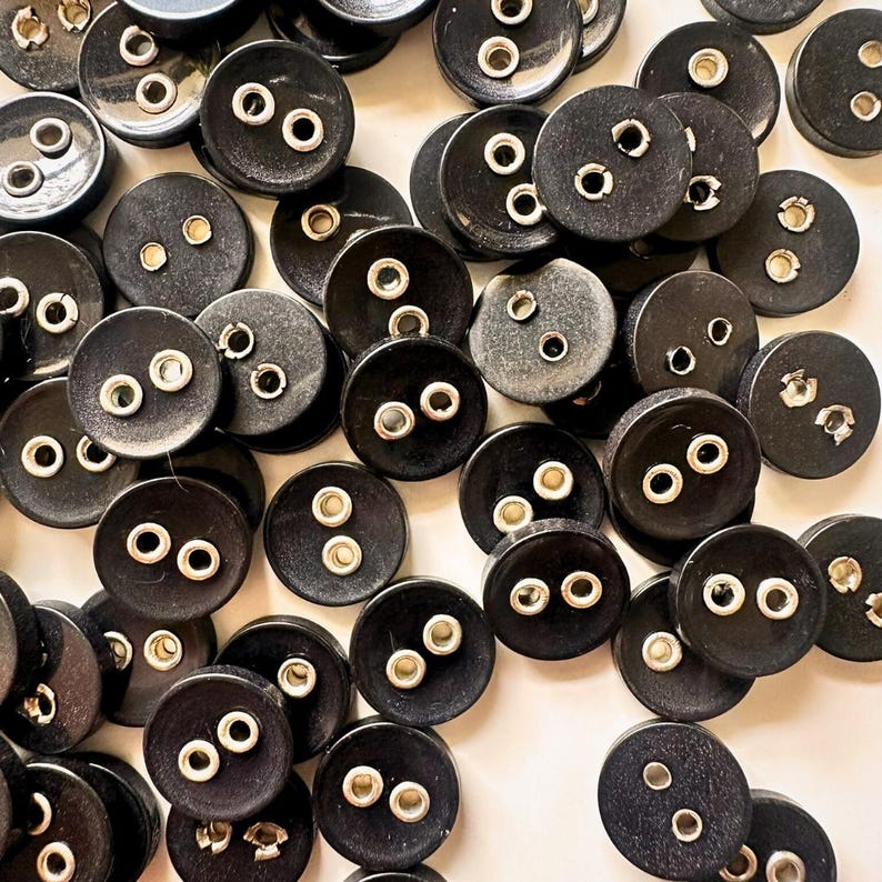 K&ouml;nnte beinhalten: Eine Ansammlung schwarzer, runder Kn&ouml;pfe mit zwei L&ouml;chern und silbernen Metall&ouml;sen. Die Kn&ouml;pfe sind auf einer hellen Oberfl&auml;che verstreut. Geeignet f&uuml;r N&auml;h- oder Bastelprojekte.