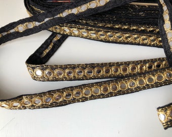 May include: Black ribbon with gold trim and small, round, reflective mirror accents. The ribbon is folded over itself in a loose coil. The gold trim is stitched around the mirrors. The ribbon is likely used for decorative purposes.
