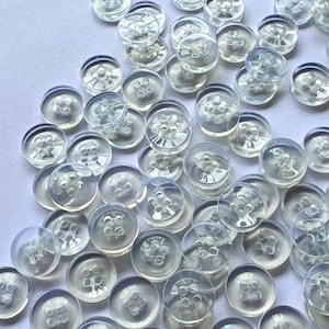 May include: A collection of numerous round, clear plastic buttons scattered across a surface. Each button features a smooth, transparent finish and four small holes in the centre for sewing, arranged in a square pattern within the circular frame.