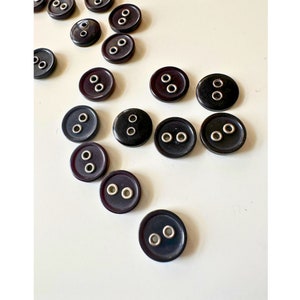 K&ouml;nnte beinhalten: Eine Sammlung dunkelbrauner, runder Kn&ouml;pfe mit zwei L&ouml;chern und silberfarbenen Metallringen. Die Kn&ouml;pfe sind auf einer wei&szlig;en Oberfl&auml;che verstreut. Ideal zum N&auml;hen, Basteln oder um Kleidungsst&uuml;cken einen klassischen Touch zu verleihen.
