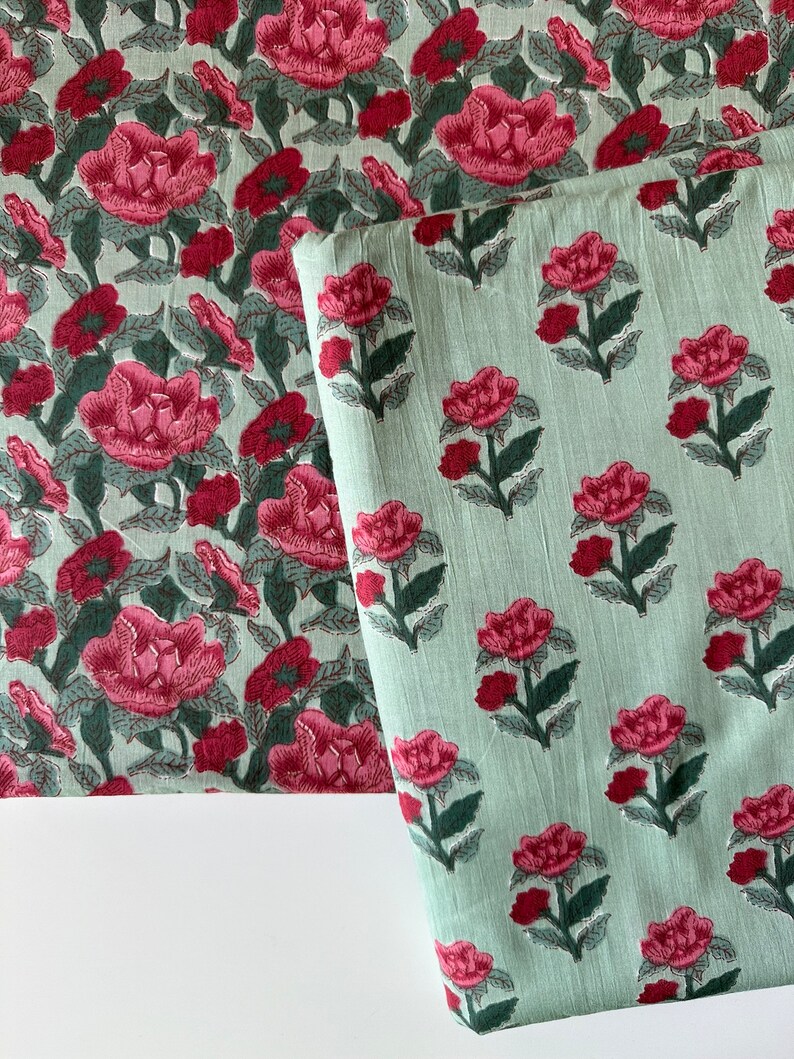 May include: Two pieces of fabric with a floral print. The top fabric is white with pink flowers and green leaves. The bottom fabric is light green with pink flowers and dark green leaves.