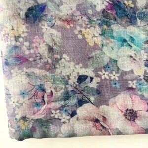 May include: A close-up of a fabric with a floral pattern. The fabric features a lavender background with a variety of flowers in shades of white, pink, blue, and yellow. The flowers are detailed and create a soft, watercolor effect.