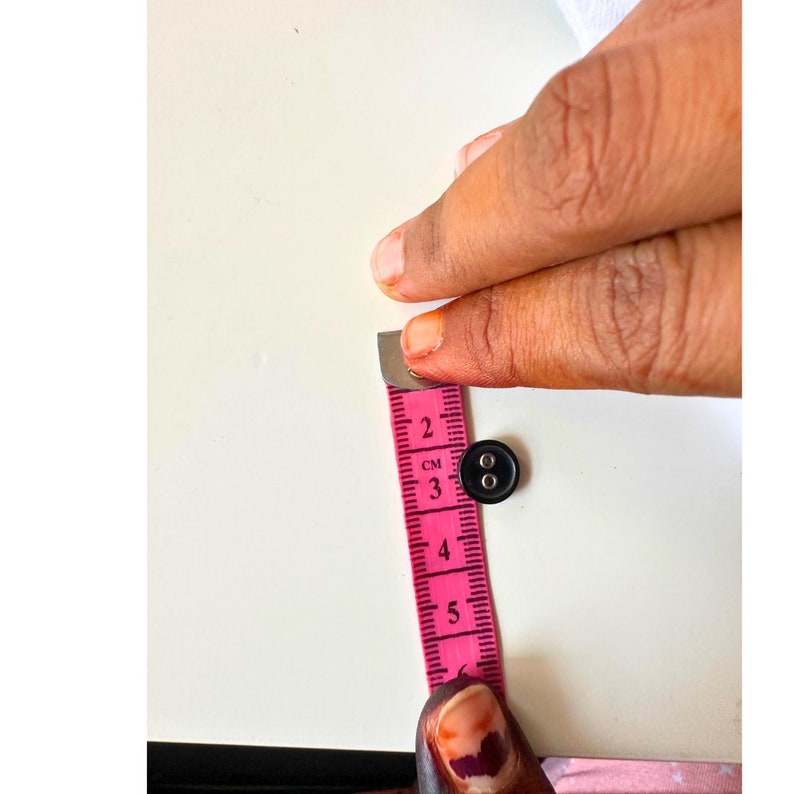 K&ouml;nnte beinhalten: Ein schwarzer Knopf neben einem rosa Ma&szlig;band. Das Band zeigt Messungen in Zentimetern an. Der Knopf hat einen Durchmesser von etwa 1,3 cm. Das Band wird von den Fingern einer Person gehalten.