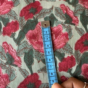 May include: A close-up of a light green fabric with a pink floral print. A blue measuring tape is placed on the fabric, showing a measurement of 9 centimeters.
