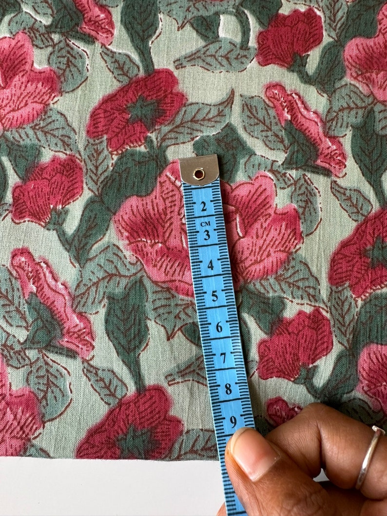 May include: A close-up of a light green fabric with a pink floral print. A blue measuring tape is placed on the fabric, showing a measurement of 9 centimeters.