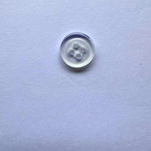 May include: A small, circular, transparent button with a smooth, glossy finish. The button features a recessed centre area containing four evenly spaced holes arranged in a square pattern for attachment to fabric or other materials.