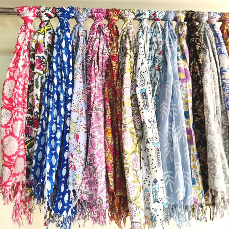 May include: A collection of colorful cotton scarves with various floral and geometric patterns. The scarves are hanging on a rack and have fringed edges.