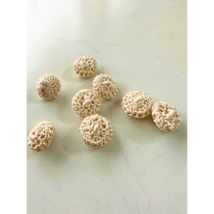 Può includere: Una collezione di bottoni all'uncinetto color crema in varie forme, tra cui rotondi e ovali. I bottoni hanno un design a trama aperta e strutturata e un piccolo bottone bianco cucito al centro. I bottoni sono sparsi su una superficie chiara.