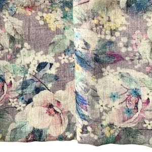 May include: Close-up of a floral fabric with a watercolor effect. The design features large, soft pink and white flowers, along with smaller blue, yellow, and white blossoms. Green leaves and stems are interspersed throughout the pattern, set against a muted purple background.