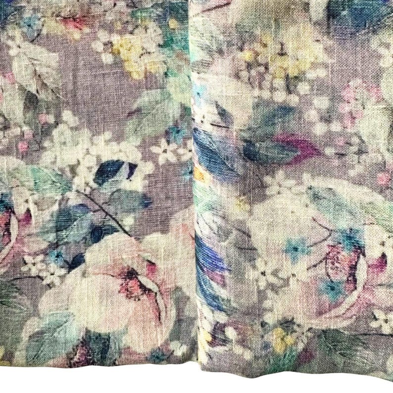 May include: Close-up of a floral fabric with a watercolor effect. The design features large, soft pink and white flowers, along with smaller blue, yellow, and white blossoms. Green leaves and stems are interspersed throughout the pattern, set against a muted purple background.