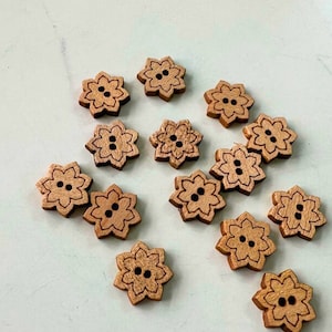 Pode incluir: Uma coleção de 14 botões de madeira em forma de flor com dois furos. Os botões são de cor castanho claro com um contorno castanho escuro e dois furos castanho escuro.