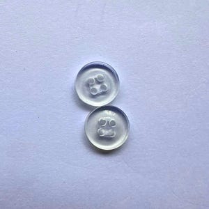 May include: Two circular, transparent buttons with a smooth, glossy finish. Each button features four small, centred holes arranged in a square pattern for attachment. The buttons are clear, allowing light to pass through their surface.