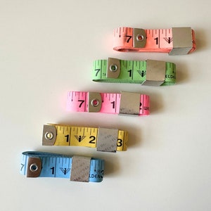 May include: Five different measuring tapes with metal clasps. The tapes are pink, green, yellow, blue, and light blue. The numbers on the tapes are black. The tapes are all in a row on a white surface.