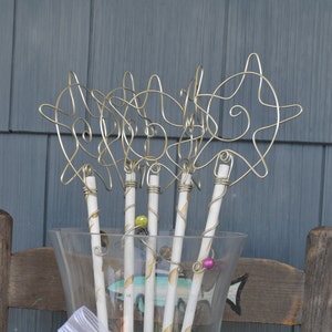 May include: A set of six gold wire decorative sticks with white painted wooden bases. The sticks are arranged in a clear glass vase. The wire designs are abstract and resemble stars or fish.