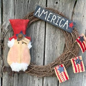 May include: A patriotic wreath with a sign that reads "AMERICA" and a red star. The wreath features a stuffed figure with a red hat and a garland of small American flags. The background is weathered wood.