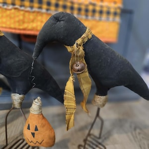 Puede incluir: Dos figuras de cuervo de tela negra sobre patas de alambre de metal. Cada cuervo tiene un pañuelo de tela a cuadros amarillo mostaza con un pequeño cascabel de metal. Un cuervo sostiene una pequeña calabaza de tela naranja con cara negra.