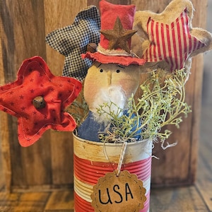 Peut inclure: Décoration patriotique avec une poupée en tissu portant un chapeau rouge et des étoiles décoratives. La poupée est dans une boîte en fer blanc rouge, blanche et bleue avec une étiquette "USA". Des étoiles décoratives rouges, noires et blanches complètent l'ensemble.