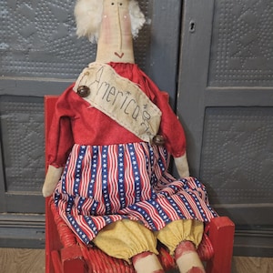 Puede incluir: Una muñeca de tela con una corona amarilla, una blusa roja de manga larga y una falda con rayas rojas, blancas y azules. Una banda beige sobre el pecho dice America 250th y tiene dos pequeñas campanas de metal sujetas a la tela.