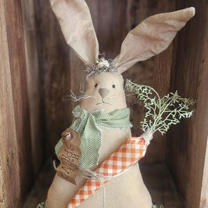 Peut inclure: Un lapin en tissu beige avec de longues oreilles, portant une écharpe verte et tenant une carotte à carreaux orange et blanc. Une étiquette en bois avec "Hello Spring" est attachée. Le lapin est décoré de verdure artificielle, parfait pour Pâques.