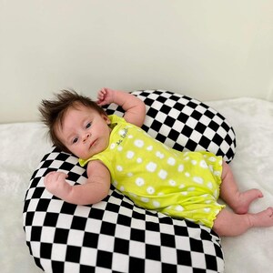Pu&ograve; includere: Un bambino con un body giallo a pois bianchi giace su un cuscino a scacchi neri e bianchi. Il body ha la scritta "so tiny" su di esso.