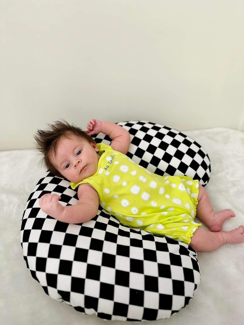 Pu&ograve; includere: Un bambino con un body giallo a pois bianchi giace su un cuscino a scacchi neri e bianchi. Il body ha la scritta "so tiny" su di esso.