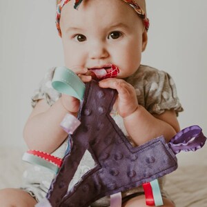 May include: A baby wearing a headband with a colorful pattern is holding a purple fabric toy with multiple colorful ribbons attached. The toy has a textured surface and is shaped like a letter.