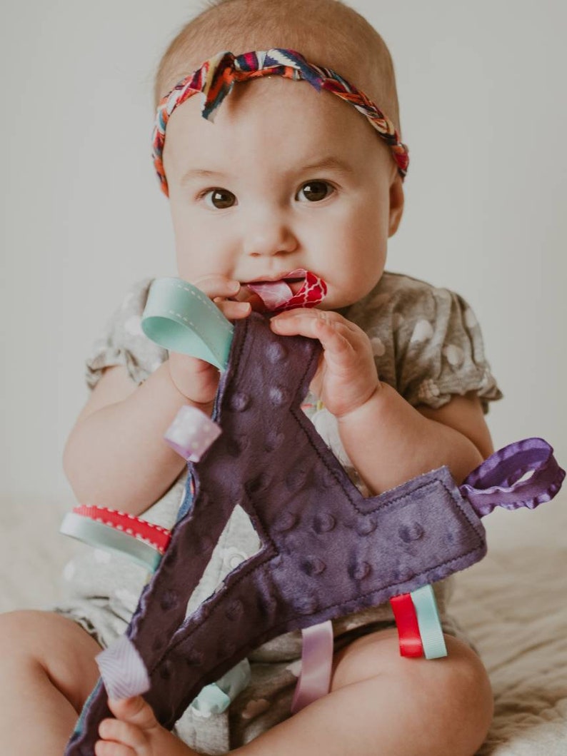 May include: A baby wearing a headband with a colorful pattern is holding a purple fabric toy with multiple colorful ribbons attached. The toy has a textured surface and is shaped like a letter.