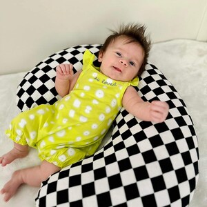 Pu&ograve; includere: Un bambino che indossa un vestito giallo con pois bianchi e il testo "so loved" sul davanti, sdraiato su un cuscino da allattamento a scacchi neri e bianchi.
