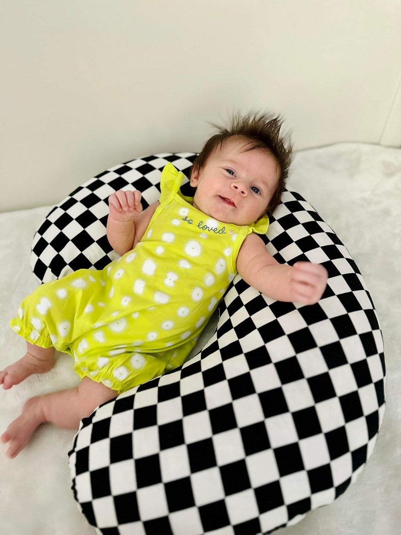Pu&ograve; includere: Un bambino che indossa un vestito giallo con pois bianchi e il testo "so loved" sul davanti, sdraiato su un cuscino da allattamento a scacchi neri e bianchi.