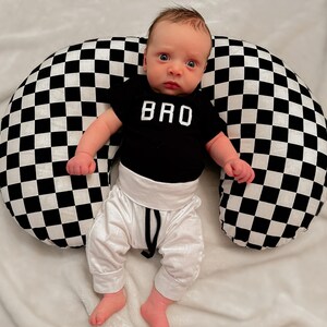Pu&ograve; includere: Un bambino che indossa un body nero con la scritta "BRO", pantaloni bianchi e un cuscino per l'allattamento a scacchi.