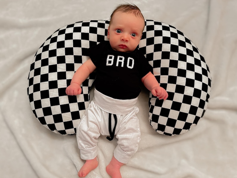 Pu&ograve; includere: Un bambino che indossa un body nero con la scritta "BRO", pantaloni bianchi e un cuscino per l'allattamento a scacchi.