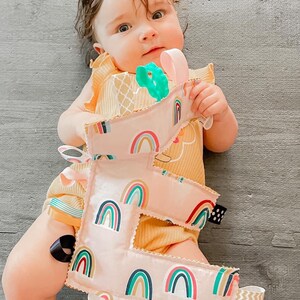 May include: A pink fabric letter E with rainbow patterns and colorful ribbons attached. The letter is held by a baby wearing a yellow onesie.