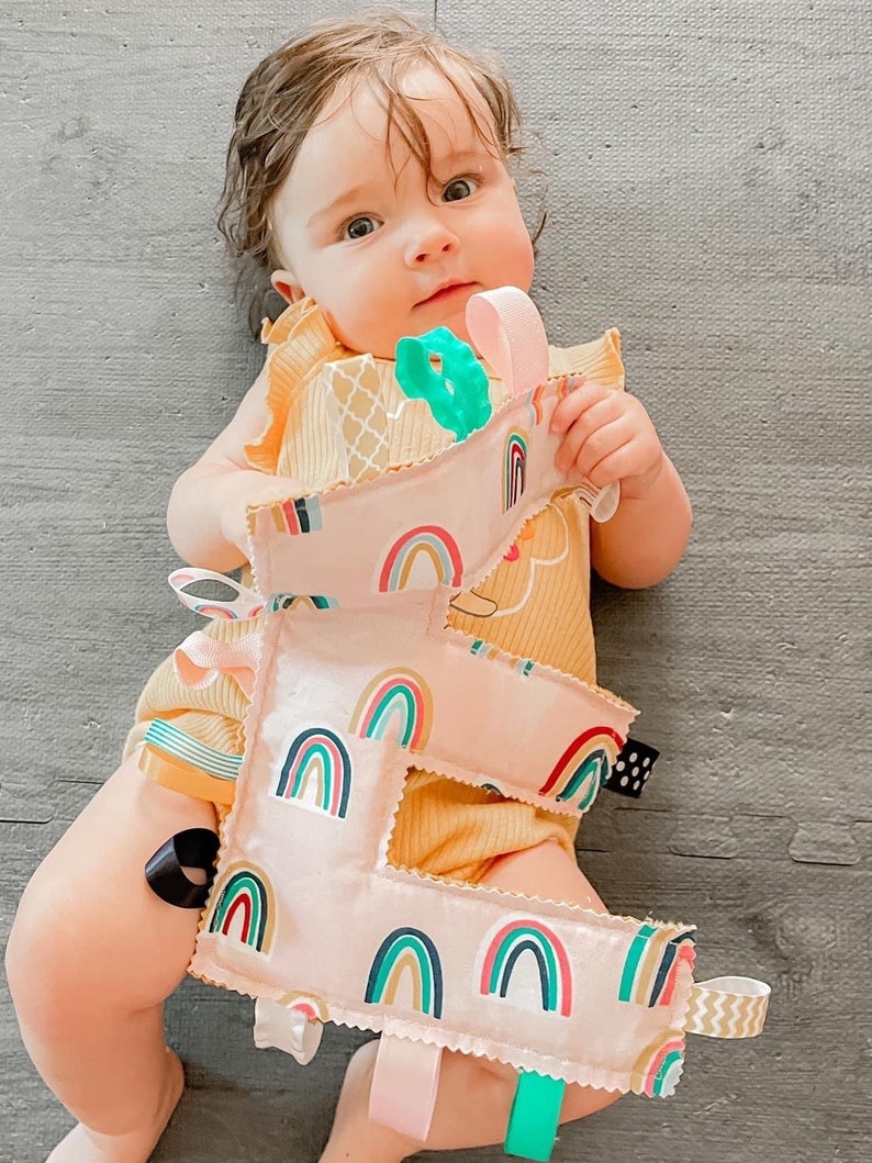May include: A pink fabric letter E with rainbow patterns and colorful ribbons attached. The letter is held by a baby wearing a yellow onesie.