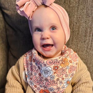 May include: A baby wearing a pink and white floral bandana bib with the text "Lil Sis" printed on it. The baby is also wearing a pink velvet headband.