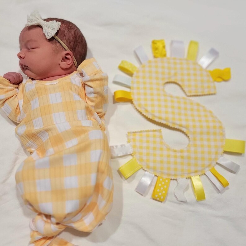 May include: A yellow and white gingham baby knotted gown with a white bow headband. The baby is lying next to a yellow and white gingham letter S with yellow ribbons.