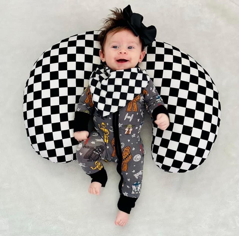 Pu&ograve; includere: Un bambino che indossa una tutina a tema Star Wars grigia e nera con un bavaglino a scacchi bianco e nero. Il bambino &egrave; seduto su un cuscino a scacchi bianco e nero.