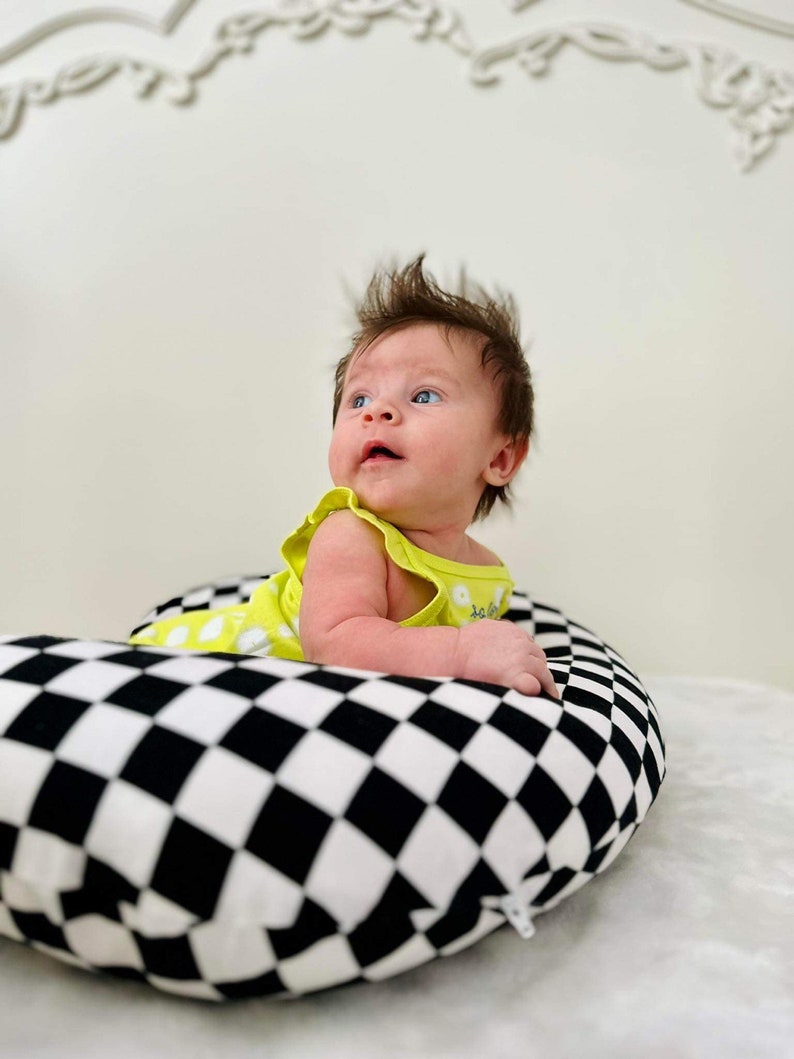 Pu&ograve; includere: Un bambino con un body giallo a pois bianchi giace su un cuscino a scacchi neri e bianchi. Il bambino sta guardando in alto e ha un'espressione sorpresa sul viso.