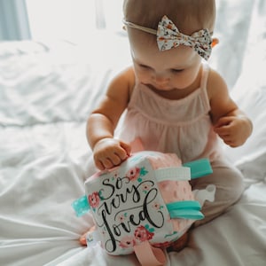 May include: A baby wearing a floral bow headband and a light pink tank top is holding a soft, pink and white cube toy. The cube has the words "So very loved" printed on it, along with floral accents and teal and pink ribbons.