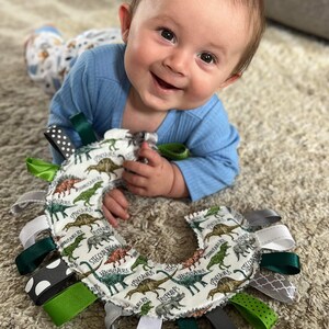 May include: A baby is playing with a dinosaur-themed sensory toy. The toy is a circle with a variety of colorful ribbons attached. The toy is made of soft fabric and has a dinosaur print.