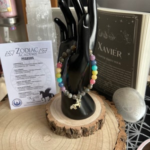 Pode incluir: Um bracelete de contas de arco-íris com um amuleto de Pégaso dourado. O bracelete está em um manequim de mão preto e uma base de madeira. O texto "Zodiac Academy Pegasus" e "Xavier Capítulo Trinta e Quatro" é visível no fundo.